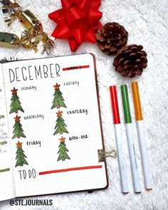 an open planner with christmas trees on it next to some pens and pineconis