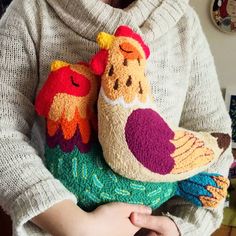 a woman holding two knitted chickens in her hands, one is wearing a sweater and the other has a bird on it