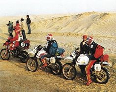 four people on dirt bikes in the desert with sand dunes behind them and one person wearing red