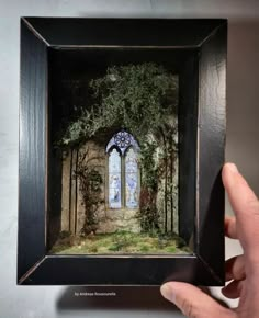 a hand holding up a miniature painting of a window in a forest with trees and grass