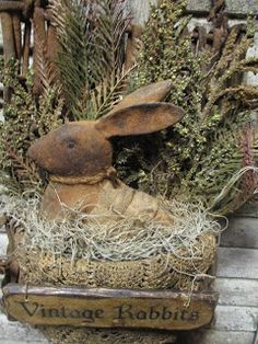 a rabbit statue sitting on top of a basket filled with grass and plants next to a wooden fence