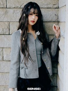 a woman standing next to a brick wall wearing a gray jacket and black skirt with her hand on her hip