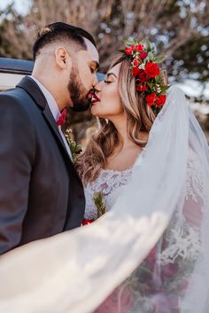 a bride and groom kissing in front of a car with red flowers on its head