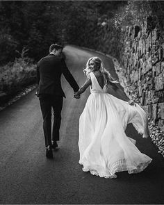 black and white photo of bride and groom holding hands walking down the road in front of a stone wall