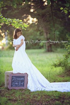 a pregnant woman in a white dress standing on a wooden box with a chalkboard sign