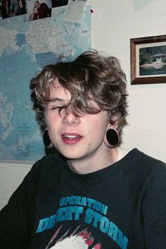 a young man with curly hair and piercings wearing a black shirt in front of a map