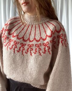 a woman is wearing a sweater with red and white designs on it, standing in front of a curtain