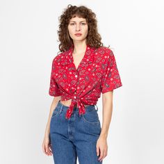 a woman standing in front of a white wall wearing jeans and a red shirt with paisley print