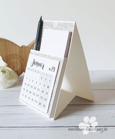 a desk calendar sitting on top of a wooden stand next to a pen and flower