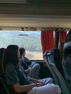 two people sitting on a bus looking out the window at mountains and trees in the distance