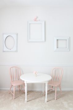 two pink chairs and a white table in a room with pictures on the wall behind them