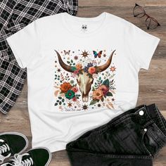 When rustic meets floral, magic happens! Our Cow Skull With Flowers tee celebrates the fusion of rugged charm and delicate blooms. Imagine a sun-kissed meadow where wildflowers twine around every curve of the skull. It's like Mother Nature decided to redecorate with a boho twist! Whether you're a flower child or just appreciate the juxtaposition, wear this tee and let your style bloom."  What's better than a soft, light cotton, quality t-shirt in your wardrobe? This comfy, contoured 100% cotton tee offers a semi-fitted silhouette and is pre-shrunk to ensure it stays a staple for longer. It features cap sleeves and a taped neck and shoulders. .: 100% cotton (fiber content may vary for different colors) .: Medium fabric (5.3 oz/yd² (180 g/m .: Semi-fitted .: Tear-away label T Shirt Butterfly, Cowgirl T Shirt, Boho T Shirt, Flower T Shirt, Skull With Flowers, Butterfly T Shirt, Boho Twists, Skull Graphic, Floral Skull