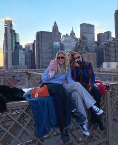 two women sitting on the edge of a bridge in front of a cityscape