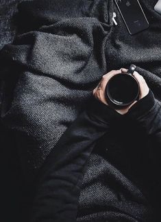 a person holding a coffee cup on top of a black blanket next to a cell phone