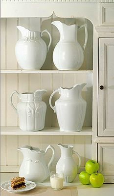 white dishes and pitchers are on shelves in a room with wood paneled walls, along with green apples