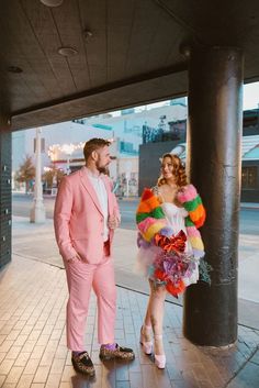 a man in a pink suit standing next to a woman wearing a colorful dress and headpiece