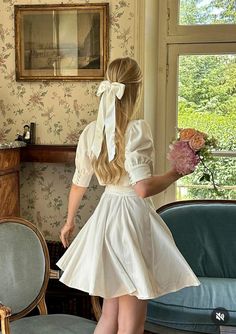 a girl in a white dress is holding flowers and looking out the window with her back to the camera