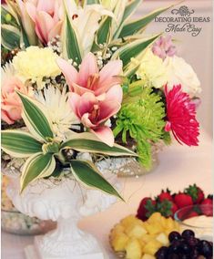 a table topped with a white vase filled with flowers and fruit next to a cake