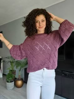 a woman with curly hair wearing a purple sweater and white pants, holding her hands on her head