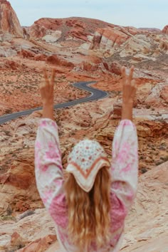 a girl with her hands up in the air on top of a rock formation looking down at a winding road