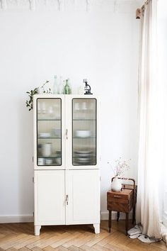 a white cabinet with two glass doors in a room next to a chair and table