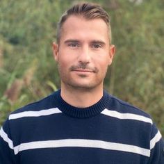 a man with a goatee standing in front of some trees and bushes wearing a striped sweater