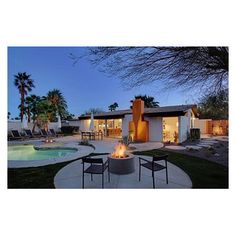 an outdoor fire pit in front of a pool at night with palm trees and chairs around it