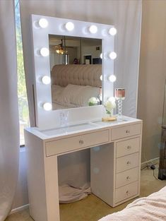 a white vanity with lights on it in front of a mirror and window, next to a bed