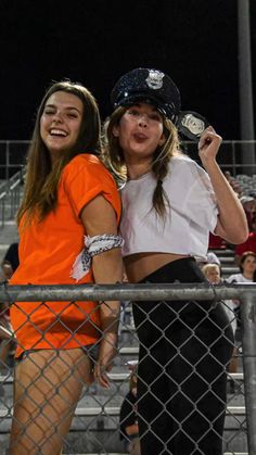 two young women standing next to each other in front of a fence at a baseball game