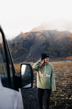 a woman is standing in front of a car talking on her cell phone while wearing a beanie