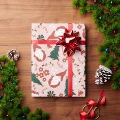 a christmas present wrapped in white paper with red ribbon and holly berries on it next to pine cones