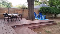 a deck with chairs and table next to a tree in a backyard area that is fenced off