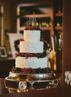 a three tiered cake with berries on top sits on a table in front of a wine rack