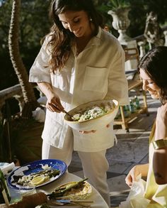 two women standing around a table with plates of food in front of them and one woman holding a bowl