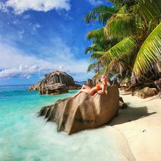 a woman laying on top of a rock next to the ocean under a palm tree