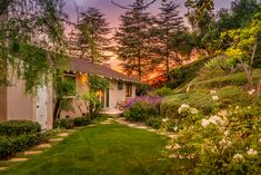 the house is surrounded by lush green grass and flowers, with trees in the background