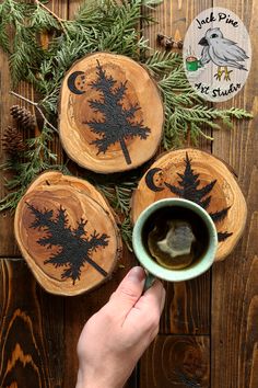 a person holding a cup of coffee in front of three wooden coasters with trees on them