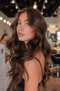 Woman with long, wavy brown hair in a well-lit salon. Rich Hair Color Ideas For Brunettes, Chocolate Brown Dark Hair, Long Hair Chocolate Brown, Chocolate Brown Hair With Light Brown Highlights, Hair Gloss For Brunettes, Glowy Brown Hair, Rich Milk Chocolate Brown Hair, Mud Brown Hair, Toner Brunette Hair