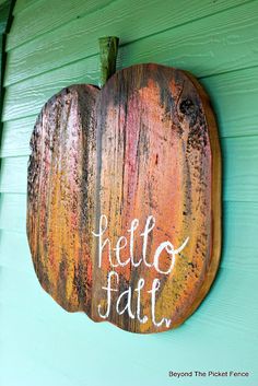 a wooden sign that says hello fall hanging on the side of a green house with an orange and yellow painted pumpkin