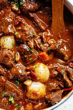 a pot full of beef stew with potatoes and carrots on the side next to a wooden spoon