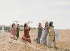 four women in long dresses are walking through the tall grass with umbrellas on their heads