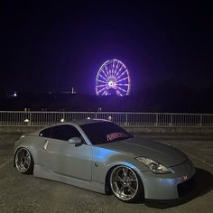 a silver sports car parked in front of a ferris wheel at night with the lights on