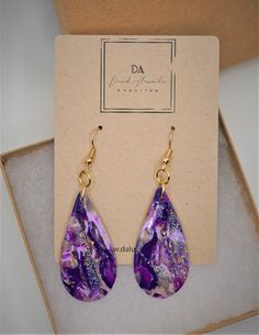 a pair of purple and gold earrings sitting on top of a cardboard box next to a card