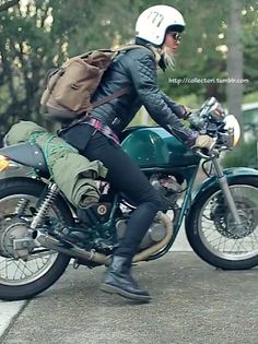 a woman riding on the back of a green motorcycle down a street with trees in the background