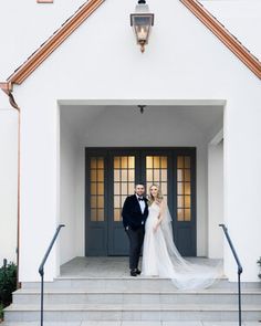 a bride and groom standing in front of a white church with blue doors, dressed in black tuxedo