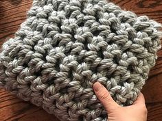 a hand is holding up a crocheted cushion on a wooden table