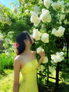 a woman in a yellow dress smelling white flowers on a tree with green grass and bushes behind her