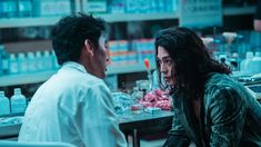 a man and woman sitting at a table in a pharmacy with bottles on the shelves