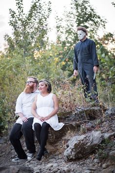 a man and woman sitting on rocks in front of trees with a scary face painted on them