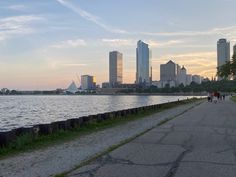 people are walking on the path next to the water in front of some tall buildings